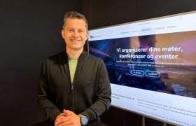 Mann med kort, mørkt hår smiler mot kamera. Han har på seg svart genser med en lys t-skjorte synlig i åpningen. En TV står på i bakgrunnen, inne på kontoret hans.