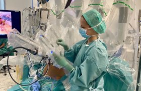 En dame står i operasjonsklær og legger plastikk på et operasjonsinstrument. I bakgrunnen ligger en pasient tildekket. Bildet er tatt i en operasjonsstue.