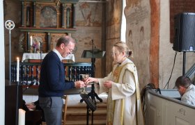 Prestgir en mann et nattverdsbeger i en gammel kirke. Presten har på seg hvit kappe med gullmønster. Mannen smiler. I bakgrunnen er et pyntet alter, malerier på veggene og tente lys. En person spiller piano til venstre, og en annen sitter bak en kirkebenk til høyre.