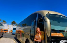 Ei dame i oransje t-skjorte og beige skjørt. Hun holder en oransje perm, og smiler. Hun står i døra til en turistbuss.