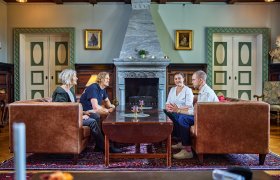 Fire personer sitter i et staselig rom i et hus. Det er en stor peis og fine, grønne dører på hver side. Det er to damer og to menn, de sitter to og to i brune skinnsofaer og prater. 