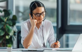 Bilde av tolk i arbeid med høretelefoner, mikrofon, skriveblokk og PC på kontorpulten