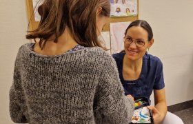 En dame med mørkt hår og briller, ikledd blå t-skjorte smiler til en ung jente med grå genser og brunt hår. Det er tegninger på veggen i bakgrunnen.