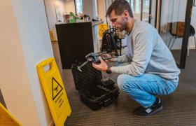 En mann med skjegg, iført olabukse og en lysegrå genser, sitter på huk og setter sammen en liten drone. Ved siden står et gult skilt med teksten "droneflygng pågår".