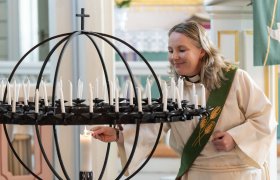 Kvinne med skulderlangt lyst hår i hvit liturgisk drakt og grønn stola tenner et lys på en stor lysglobe i en kirke.