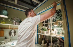 En maler står på et kjøkken og strekker seg til toppen av en list. Listen ligger rundt et vindu, og er maskert med blå teip.