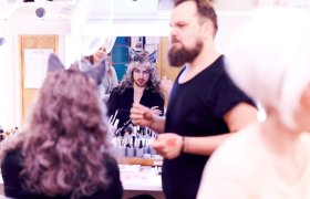 Et rom fylt med sminkeutstyr og speil. En person med langt, bølgete hår og dyreører får sminke påført. En annen person står i bakgrunnen, mens en tredje person med skjegg og svart t-skjorte holder en sminkebørste.