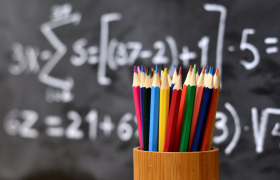 I forgrunnen er det en orange kopp med fargeblyanter, i bakgrunnen er det en tavle med matematiske formler på.