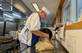 En mann kledd i hvitt heller en stor deig utover en metallbenk. 