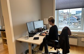 En kvinne sitter ved et skrivebord på kontor og jobber på to dataskjermer. Hun har mørk overdel og lyse jeans, og på stolen henger en jakke. Utenfor vinduet ser man en bygate med bygninger og mennesker.