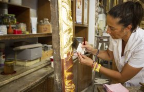 En dame sitter og forgyller et kunstverk på verkstedet sitt.