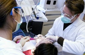 Tannlege polerer tennene til en pasient, mens en tannhelsesekretær holder et beger med slipepasta som brukes i poleringen.