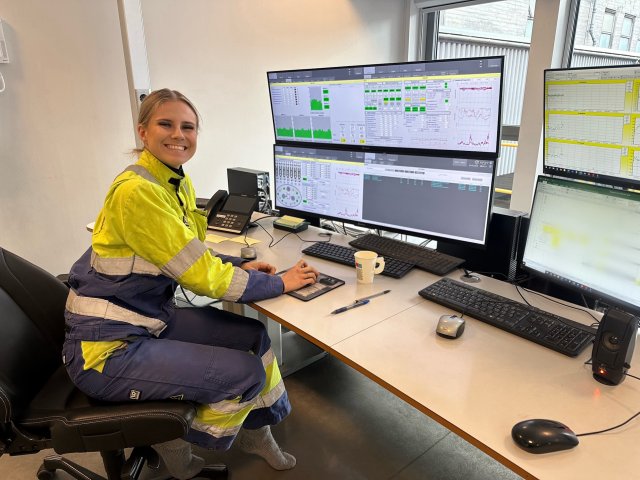 Dame med lyst hår smiler mot kamera. Hun sitter ved et skrivebord med fire store pc-skjermer foran seg.