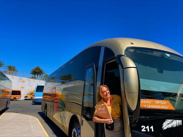 Ei dame i oransje t-skjorte og beige skjørt. Hun holder en oransje perm, og smiler. Hun står i døra til en turistbuss.