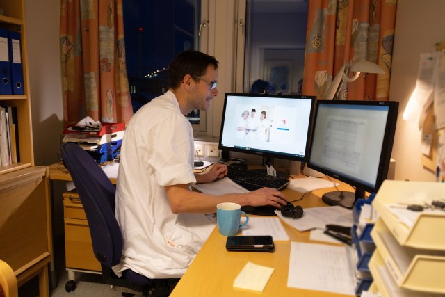 En mann i hvite sykehusklær og briller. Han jobber foran to pc-skjermer. Han er på et kontor i sykehuset.
