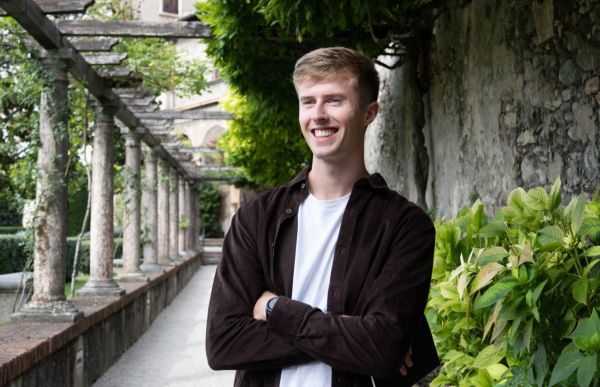 En ung mann står med armene i kors langs en steinmur omgitt av grønne planter. 